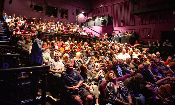 The camera faces an auditorium full of audience members, illuminated by theatre lighting. A man in a denim shirt and hat stands on the steps to the si